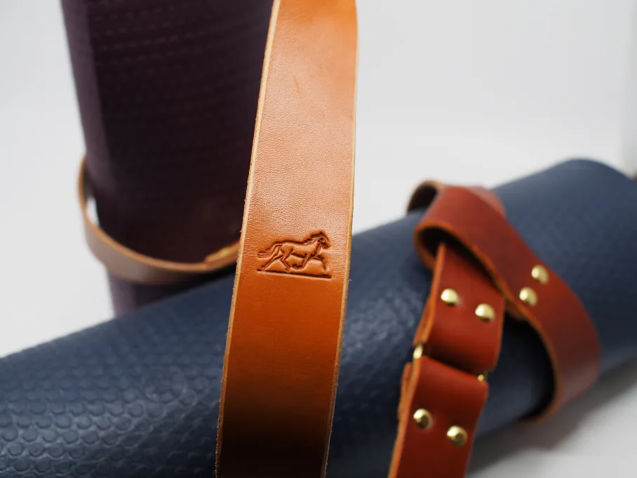the trotting horse stands out on the tan shoulder strap of a yoga sling on a purple yoga met; shiny brass rivets and a brass ring hold a looped chestnut strap around a navy blue yoga mat in the slightly blurry backgorund